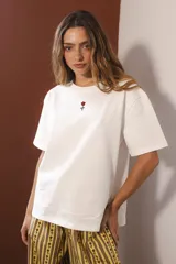 Remera blanca de algodón con cuello redondo y manga corta. Presenta un pequeño bordado de una flor roja en el centro del pecho.