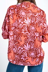 Blusa oversize de poliéster con estampado de animales y plantas en tonos rosa y marrón. Tiene cuello camisero, mangas largas y cierre frontal con lazos.