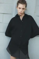Camisa negra de cuello solapa y manga larga amplia. Tiene un bolsillo de parche en el delantero, detalle de pliegues y cierre frontal con botones.