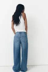 Pantalón de jean celeste de tiro alto, corte wide leg y largo hasta el tobillo. Presenta un diseño asimétrico en la cintura con cierre frontal cruzado con cremallera y botones metálicos.