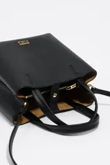 Bolso pequeño tipo tote o cubo de piel vacuna texturizada color negro, con asas cortas superiores y correa larga ajustable para llevar al hombro o cruzado. Presenta el logo metálico de Bimba y Lola en dorado en el frente.