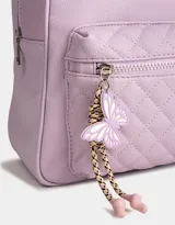 Mochila lila de cuerina con efecto acolchado y colgante de mariposa. Tiene correas regulables, manija superior, acceso con cierre superior y bolsillo frontal con cierre y tirador de goma en forma de mariposa.