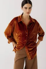Camisa color marrón de terciopelo, corte holgado, con cuello clásico, cierre frontal con botones, mangas largas con puños abotonados, dos bolsillos de parche en el pecho y ruedo recto.