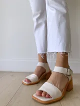 Sandalias de cuero color crudo, con tira ancha sobre los dedos, pulsera al tobillo con hebilla metálica y taco de madera de 6 cm de altura.