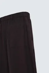 Pantalón marrón oscuro de corte ancho, confeccionado en mezcla de algodón y lino. Presenta cintura elástica, bolsillos laterales y traseros, pliegues frontales y cierre con cremallera y botón.
