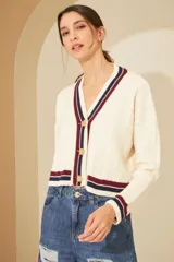 Cardigan color crema de tejido de punto con trenzas y aranes, con cuello en V y botones dorados. Presenta ribetes a rayas en azul marino y bordó en el cuello, puños y ruedo.