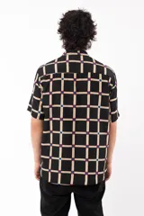 Camisa de manga corta con calce relajado y cuello tipo sastre, confeccionada en rayón. Presenta un diseño de cuadrícula beige sobre fondo negro con detalles en azul y rojo en las intersecciones.