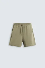 Bermuda color verde oliva de corte relaxed fit, confeccionada en tejido interlock de algodón con elasticidad. Tiene cintura elástica ajustable con cordón, bolsillos laterales y bolsillo trasero de parche.