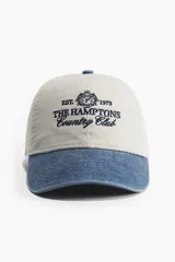 Gorra de béisbol color beige con visera azul denim, bordado frontal con el texto "Est. 1973 The Hamptons Country Club" y un escudo.
