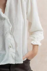 Blusa color blanco de lino con cuello redondo con volados, abotonada al frente y mangas largas con elástico en los puños.