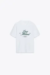Camiseta blanca de algodón con cuello redondo y mangas cortas. Estampado frontal con el texto "Luce Milano Est. 1975" en color verde oscuro.