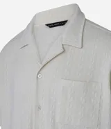 Camisa de jacquard color crema, de corte oversized, con cuello solapa, manga corta, abotonadura frontal, bolsillo en el pecho y textura en relieve.