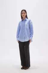 Camisa oversize de poliéster, con cuello camisero y cierre frontal de botones. Presenta un diseño de rayas verticales azules y blancas, con bloques de rayas horizontales en los puños y el ruedo inferior.