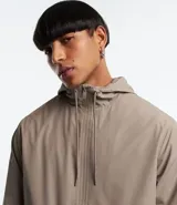 Campera rompevientos color verde oliva, con capucha ajustable con cordón, cierre frontal con cremallera, bolsillos laterales con cremallera y puños elásticos.