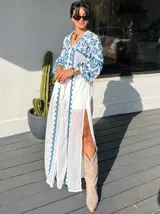Vestido largo blanco con bordados azules, escote en V y mangas largas abullonadas. Presenta una abertura lateral alta.