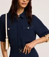 Camisa color beige de tela airflow, con cuello camisero, mangas cortas con detalle de pliegue, bolsillos con solapa en el pecho y cierre frontal con botones metalizados.