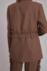 Blazer marrón con solapas de muesca, cierre frontal con lazo y mangas largas. Presenta volados a lo largo de las costuras laterales del pantalón a juego.