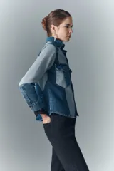 Campera recta de denim rígido con combinación de recortes en diferentes tonos de azul, cuello camisero, cierre frontal con botones metálicos, bolsillos con solapa y botones metálicos en el pecho, y puños con detalle de encaje negro.