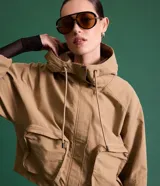 Campera corta color marrón claro, confeccionada en sarga de algodón y poliamida, con cuello alto, capucha y bolsillos delanteros con solapa. Tiene cierre frontal con botones a presión y mangas largas con puños elásticos.