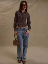 Camisa de manga larga color marrón oscuro con finas rayas verticales blancas. Presenta un corte entallado y ligeramente cropped, con cuello camisero y cierre frontal de botones.