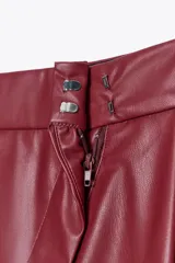 Pantalón de cuero sintético color bordó, de tiro medio, corte recto y cintura cruzada con trabillas. Cuenta con bolsillos laterales, pinzas delanteras y cierre frontal con cremallera, botón y gancho metálico.