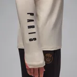 Remera de manga larga color crema con detalles en salmón en los hombros, con logo de Jordan y escudo del Paris Saint-Germain en el pecho y leyenda "PARIS" estampada verticalmente en la manga izquierda.