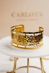 Brazalete ancho dorado con textura irregular y aberturas rectangulares en los bordes.