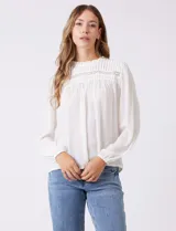 Blusa blanca de gasa plisada con encaje calado en el cuello y el pecho. Cuello a la base fruncido, manga larga con puño elastizado y espalda lisa con abertura tipo gota y botón.