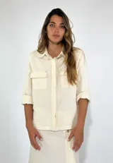 Chaqueta color crudo con cuello camisero, cierre frontal con botones, mangas 3/4 con puños enrollados y dos bolsillos de parche en el pecho.