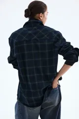 Camisa oversized de franela suave con estampado a cuadros en tonos azul marino, verde oscuro y beige. Presenta cuello, botonadura frontal con botones dorados, canesú en la espalda, hombros caídos, bolsillos superiores con solapa y botón dorado, y puños abotonados. El bajo es ligeramente redondeado.