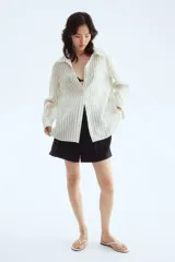 Camisa de lino color blanco con rayas verticales negras, cuello clásico, cierre frontal con botones, manga larga con puños abotonados, bolsillo de parche en el pecho y bajo redondeado.