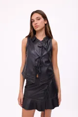 Falda corta negra de efecto cuero, estilo wrap o cruzada, con ajuste lateral mediante lazos de cuero y terminación con volante o ruedo con volado. Presenta costuras contrastantes en color claro.