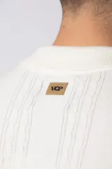 Camisa tejida color crudo, de manga corta, con cuello bowling y botones frontales. Presenta un tejido con textura de trenzas verticales y rib negro en los puños.