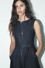 Vestido midi de denim azul oscuro, sin mangas, con cuello redondo, bolsillos laterales, cinturón con hebilla metálica y cierre frontal con cremallera.