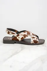 Sandalias bajas con tiras cruzadas de cuero con estampado de vaca en tonos marrón y blanco. Tira trasera con hebilla dorada y suela plana color marrón.