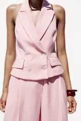 Chaleco rosa con cuello halter y escote pico con solapa. Espalda descubierta. Bolsillos delanteros con solapa. Cierre frontal cruzado con botones.