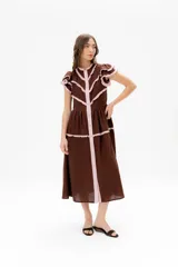 Vestido midi marrón de lino con cuello mao y cierre frontal de botones. Presenta detalles de volados en las mangas cortas y ribetes en color rosado pálido que delinean el frente y los hombros.