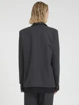 Blazer de mujer color gris oscuro con un sutil patrón de cuadros. Presenta solapas clásicas, cierre frontal de dos botones y bolsillos con tapa. Se combina con un top negro de encaje y pantalones a juego.