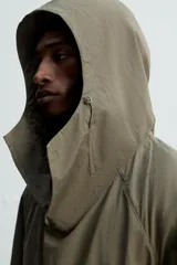 Parka color verde militar de corte amplio, confeccionada en tejido técnico ligero. Cuenta con cuello alto con capucha ajustable, cierre frontal con botones, mangas largas con puños ajustables, bolsillos de plastrón con solapa en la parte delantera, cintura ajustable con cordón y bajo con aberturas laterales ajustables con cordón.