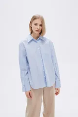 Camisa celeste con finas rayas verticales blancas, de corte holgado, manga larga con puños abotonados y bolsillo de parche en el pecho.