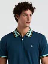 Chomba azul petróleo de manga corta con cuello polo y puños acanalados a rayas en celeste, naranja y blanco. Logo bordado en el pecho. Cierre con dos botones.