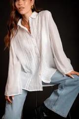 Camisa color beige claro, de corte holgado, con cuello clásico, cierre frontal con botones, mangas largas con puños abotonados y detalles de broderie en la parte delantera.