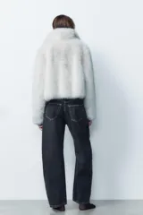 Abrigo corto color blanco de efecto piel, con cuello solapa y manga larga. Tiene bolsillos laterales ocultos en la costura, forro interior combinado y cierre frontal con ganchos metálicos forrados.