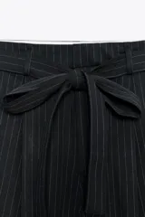 Pantalón negro con finas rayas verticales blancas, de tiro alto y corte ancho. Incluye cinturón de lazo en el mismo tejido, bolsillos laterales y pinzas.