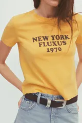 Camiseta amarilla de algodón con cuello redondo, manga corta y estampado marrón con el texto "New York Fluxus 1970".