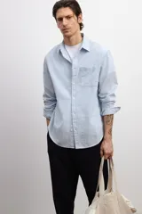 Camisa de hombre de corte holgado, confeccionada en tela de sirsaca a rayas verticales celestes y blancas. Tiene cuello inglés, tapeta clásica con botones, bolsillo de parche en el pecho, mangas largas con puños abotonados y bajo redondeado.
