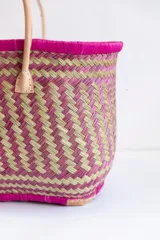 Bolso tipo tote de raffia natural con diseño tejido, interior forrado en tela fucsia y doble asa de cuero color beige. Tiene base rectangular y detalles de cuero en las esquinas inferiores.
