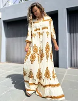 Vestido largo estilo túnica o camisero, color crudo con estampado en tono marrón/beige. Presenta cuello en V con botones frontales y mangas tres cuartos. El diseño es holgado, con varios niveles de volados en la falda y un estampado étnico o tribal repetitivo.