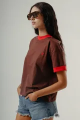 Camiseta de manga corta color marrón con cuello redondo y puños en contraste de color rojo. Presenta un pequeño bordado de una letra 'R' en color rojo en el pecho.