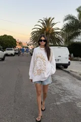 Buzo de tejido de punto color crudo, con cuello redondo y manga larga. Presenta un bordado frontal grande con un diseño abstracto que simula un rostro con rayos solares.